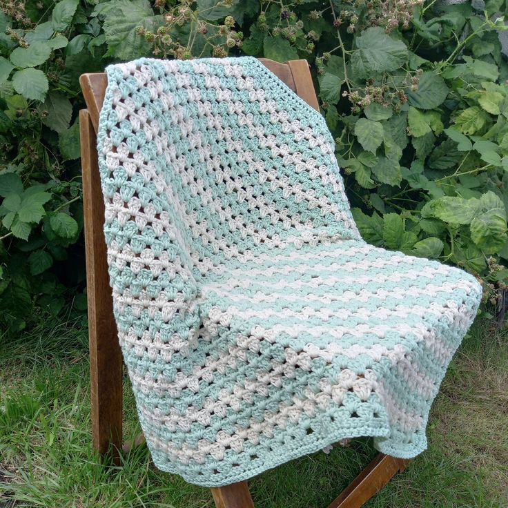 a crocheted blanket sitting on top of a wooden chair in front of bushes