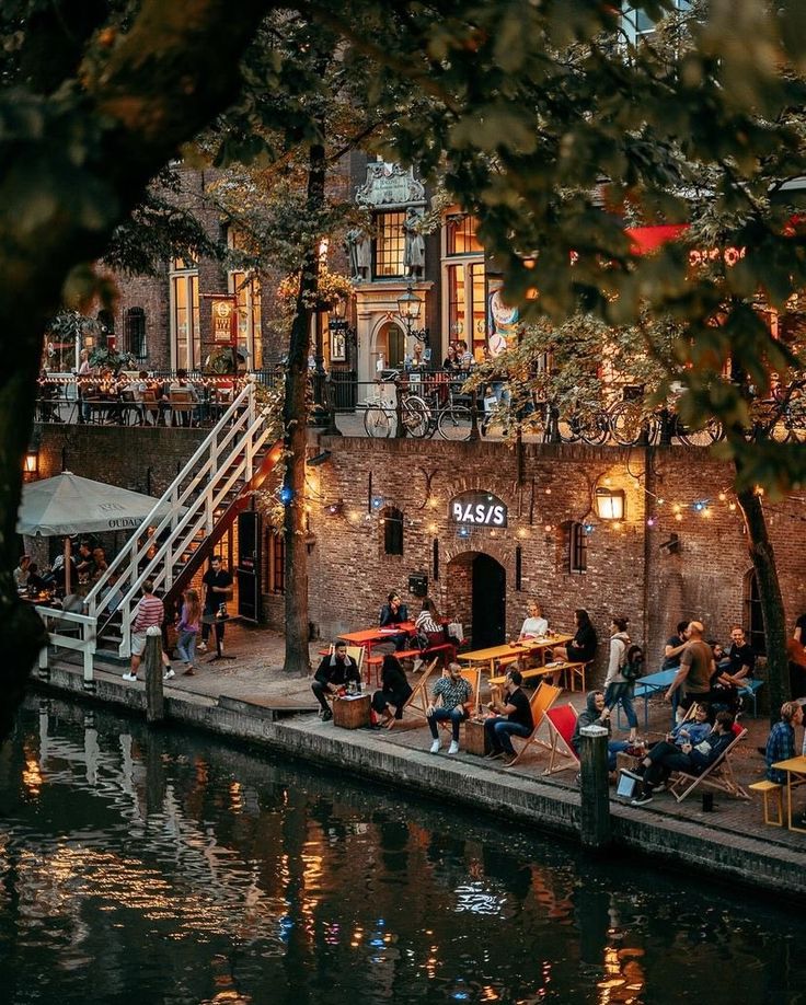 people sitting at tables on the side of a river in front of an old brick building