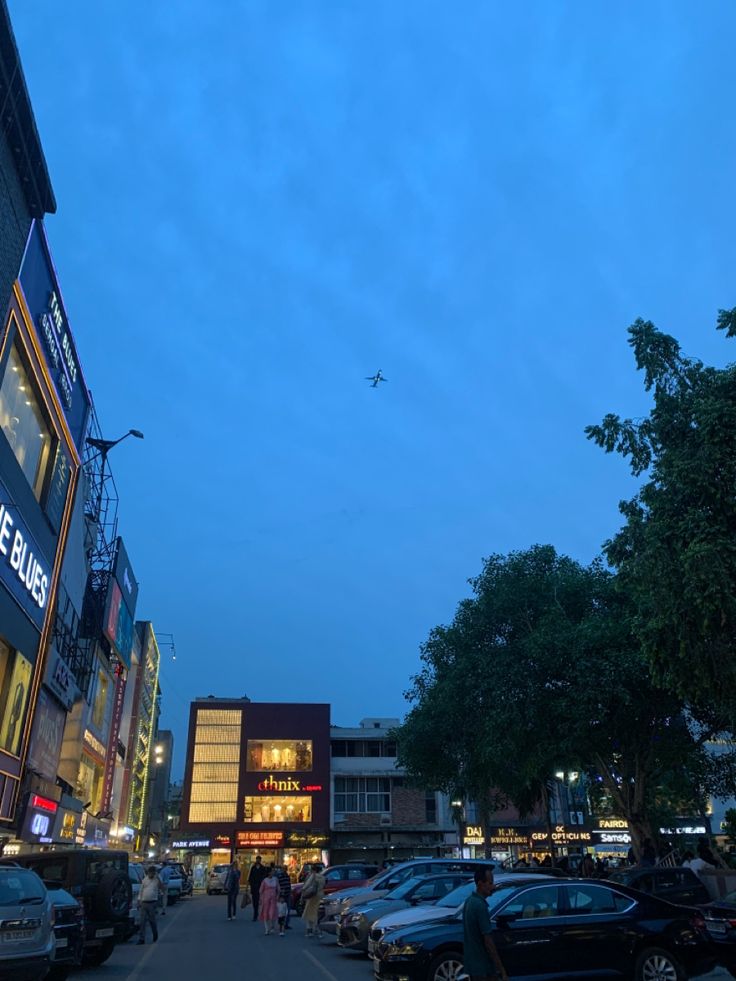 cars parked on the side of a street next to tall buildings at dusk with an airplane flying overhead