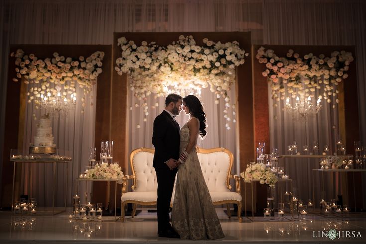 a bride and groom standing in front of a stage with flowers on the wall behind them