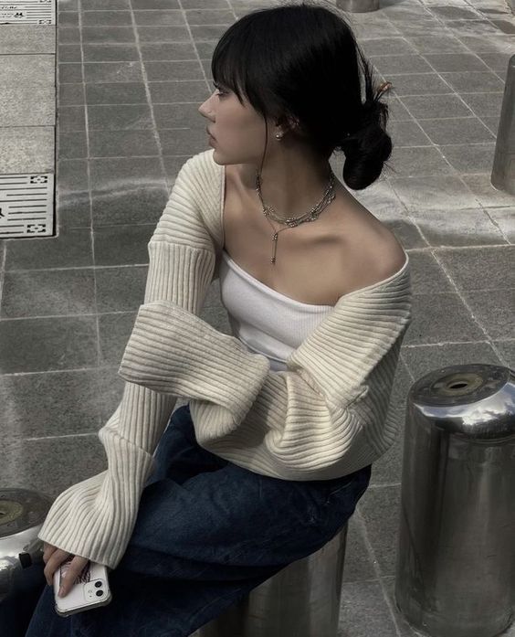 a woman sitting on top of a metal pole next to a trash can wearing a white sweater