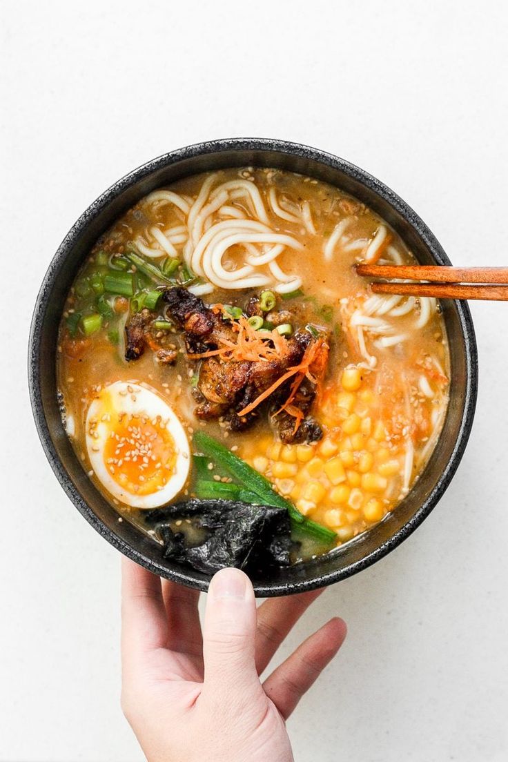 someone holding a bowl of ramen with noodles, carrots and an egg