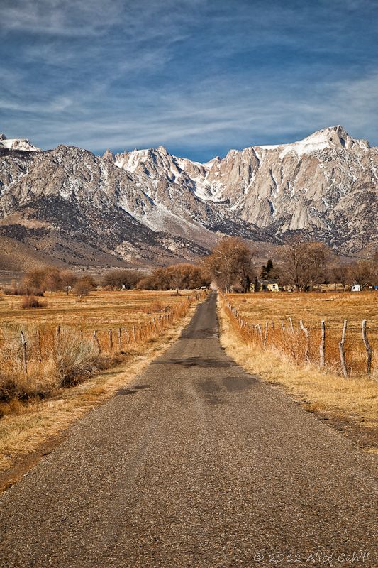 an empty dirt road in front of mountains with snow on the top and brown grass to the side