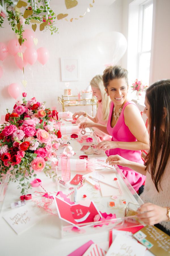 three women are sitting at a table with pink decorations and flowers on it, one woman is cutting paper