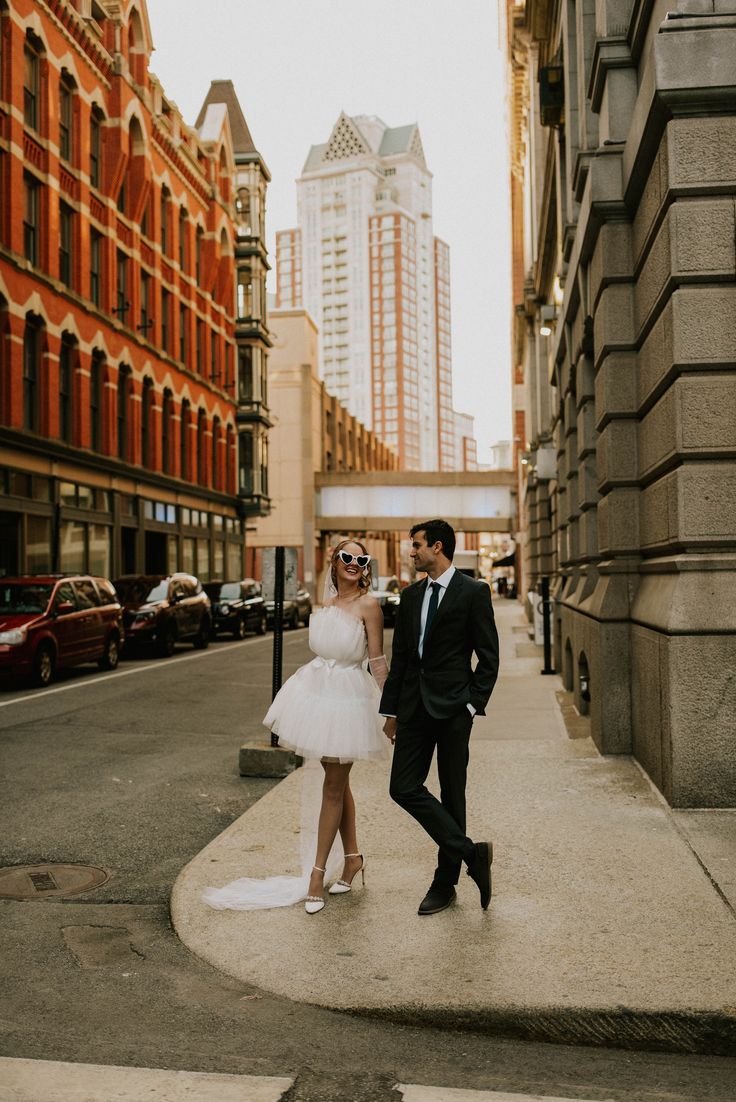 a man and woman are standing on the sidewalk in front of tall buildings, dressed in white