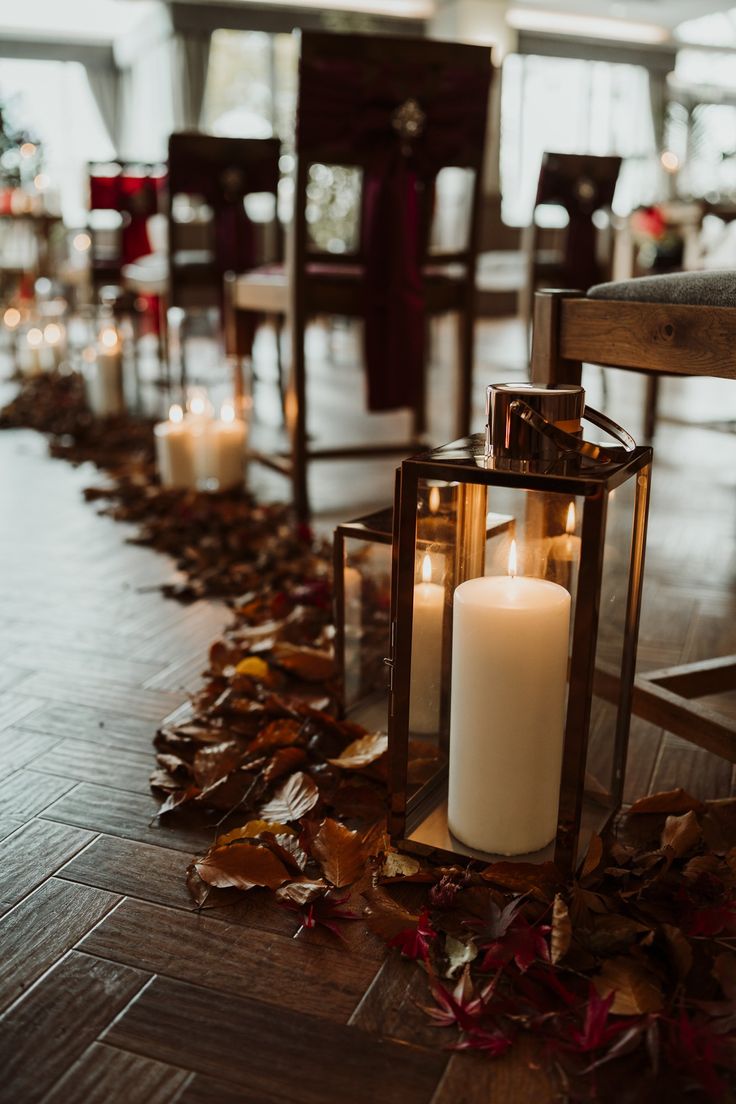 two lit candles sitting on top of a wooden floor next to some chairs and fall leaves