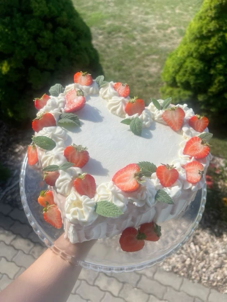 a person holding up a cake with strawberries on it in front of some bushes