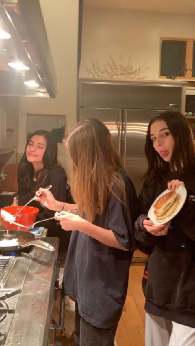 three girls standing in a kitchen holding plates with hotdogs on them and one girl eating