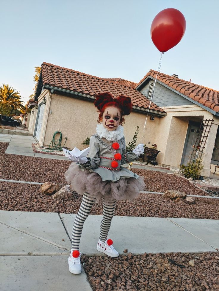 a creepy clown is standing on the sidewalk in front of a house with a red balloon