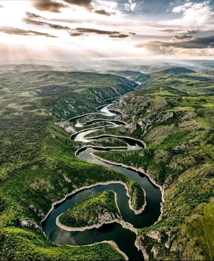 an aerial view of a river surrounded by green hills and trees with sun rays coming through the clouds