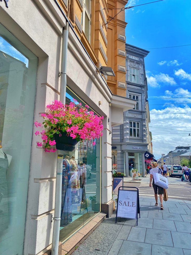 people walking down the sidewalk in front of a store with pink flowers hanging from it's windows