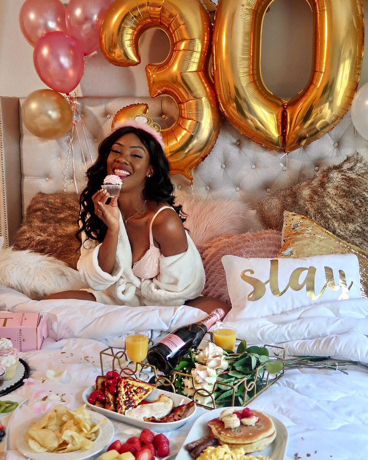 a woman sitting on a bed with balloons and food in front of her, smiling