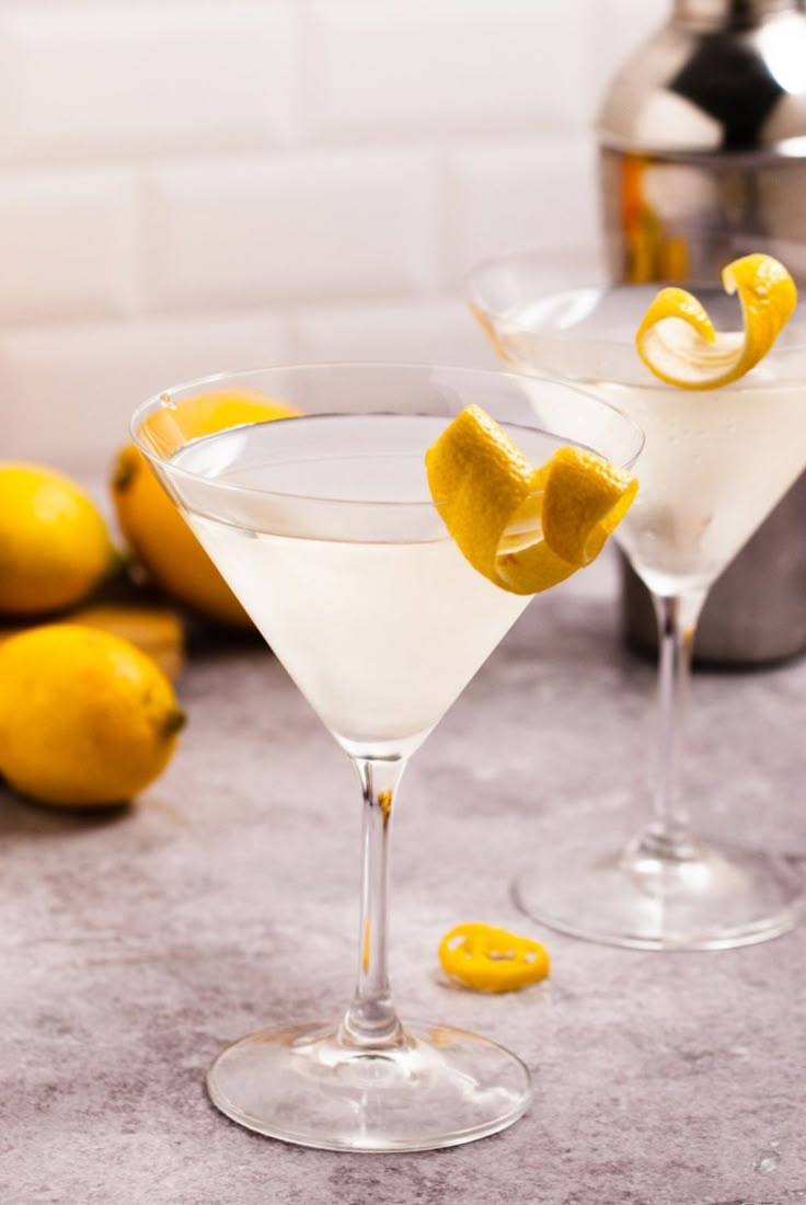 two martinis with lemon peel garnish sit on a counter next to some lemons