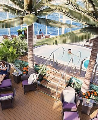 an outdoor swimming pool with lounge chairs and palm trees in the foreground, people sitting at tables next to it