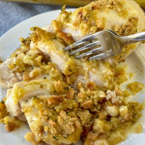 a white plate topped with chicken covered in stuffing next to a fork and bread roll