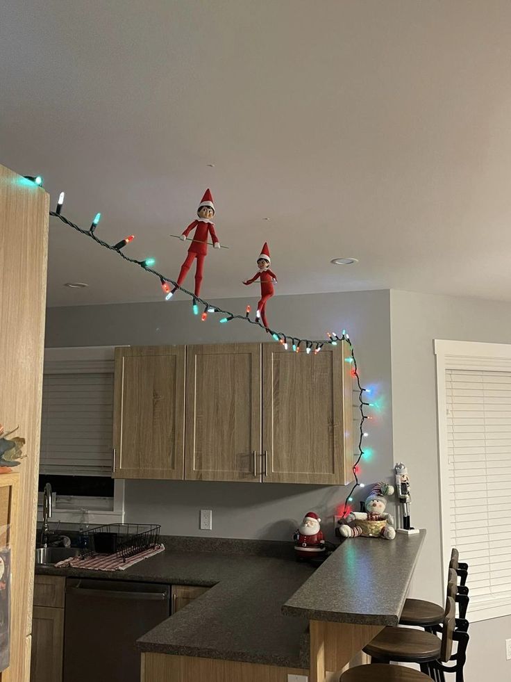 two elfs hanging from the ceiling in a kitchen with christmas lights strung across the room