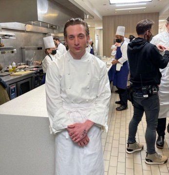 a man standing next to a counter in a kitchen with other people working behind him