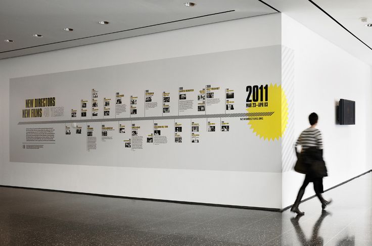 a woman walking past a white wall with yellow and black pictures on the wall next to it