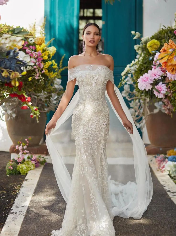 a woman in an off the shoulder wedding dress standing on a sidewalk with flowers behind her