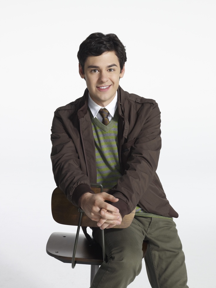 a man sitting on top of a chair wearing a brown jacket and green sweater with his hands in his pockets