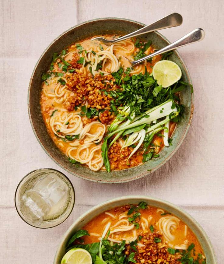 two bowls filled with noodles and greens on top of a white table cloth next to silver spoons