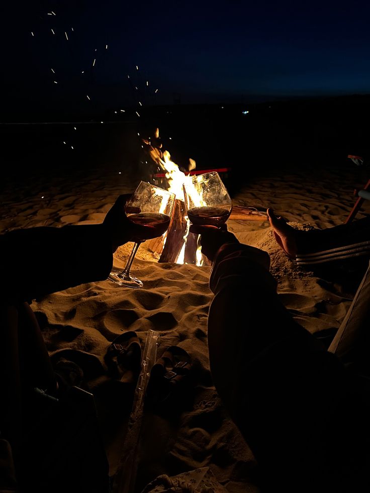 people sitting around a campfire with wine glasses in front of them at night on the beach