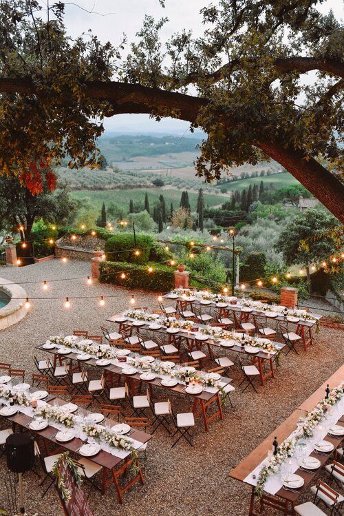 an outdoor dining area with tables and chairs set up for dinner under a tree, surrounded by greenery