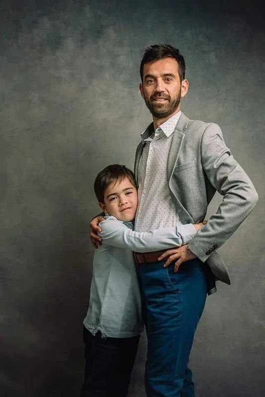 a man standing next to a little boy in front of a gray wall with his arms around him