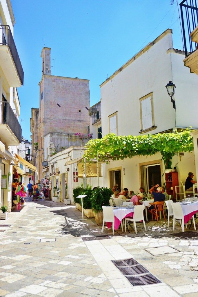 people are sitting at tables in the middle of an alleyway with white buildings and greenery on either side