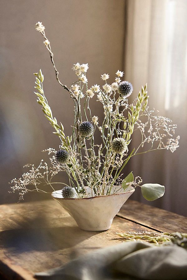 an arrangement of flowers in a white bowl on a wooden table next to a window