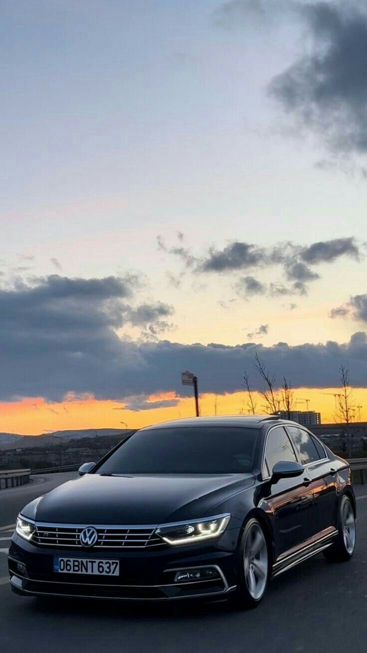 a black car is driving down the road at sunset with clouds in the sky behind it