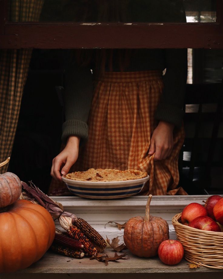 a person holding a pie in front of some pumpkins and other autumn decorations on a window sill