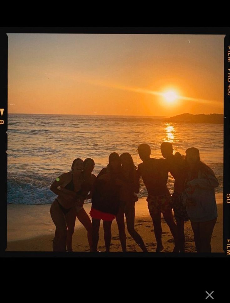 a group of people standing on top of a beach next to the ocean at sunset