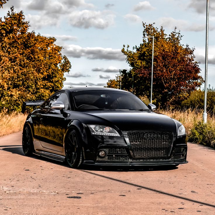 a black car parked on the side of a road next to tall grass and trees