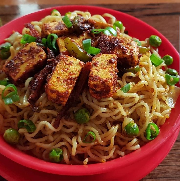 a red bowl filled with noodles and tofu on top of a wooden table next to chopsticks