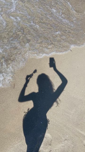 a shadow of a woman on the beach with a cell phone and bottle in her hand