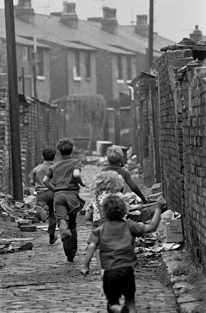 black and white photograph of children running down the street in front of brick buildings with trash on the ground