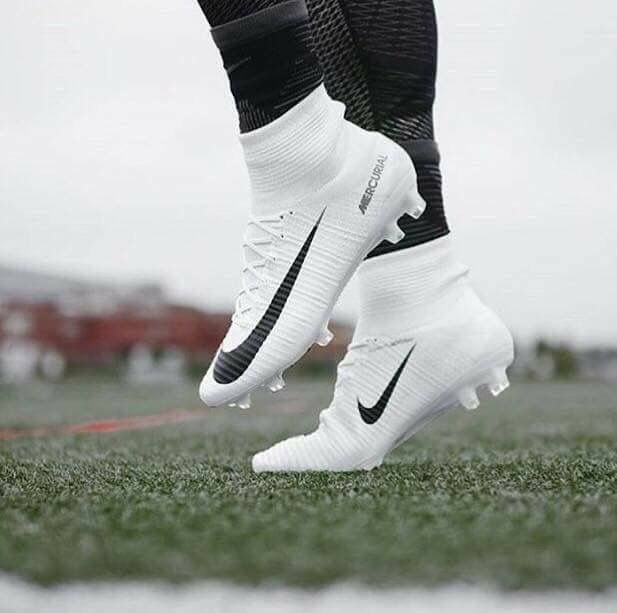 the feet of a soccer player in white and black shoes on grass with buildings in the background