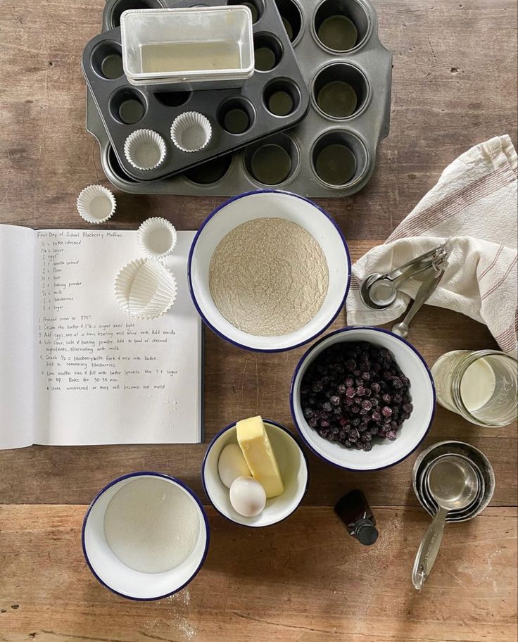 an open book sitting on top of a wooden table next to bowls and pans