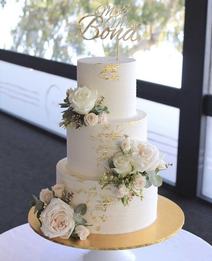 a three tiered wedding cake with white flowers and gold trimmings on a table