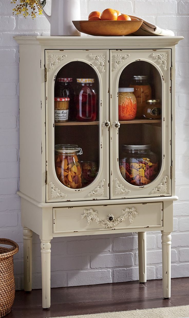 a white cabinet with glass doors holding jars and fruit