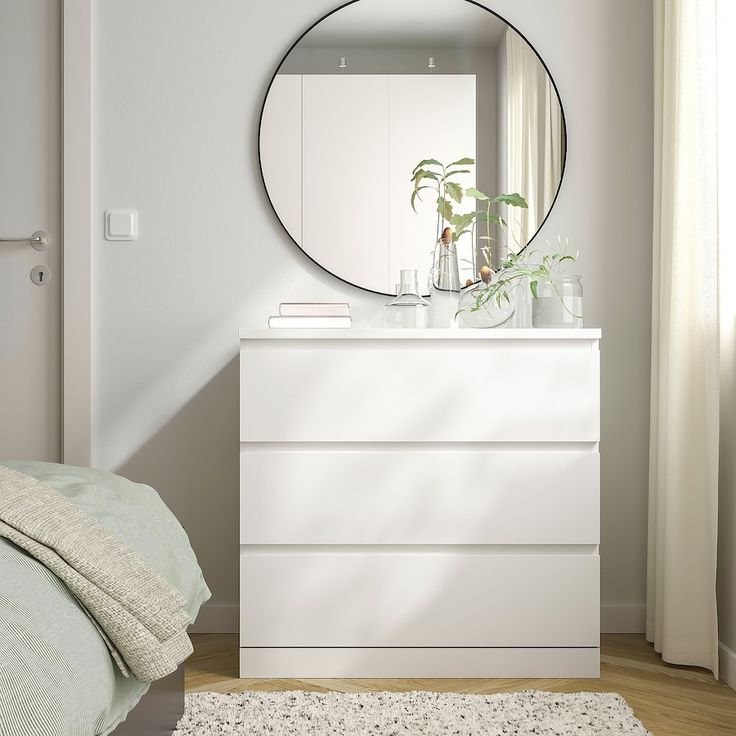 a white dresser sitting next to a bed under a round mirror