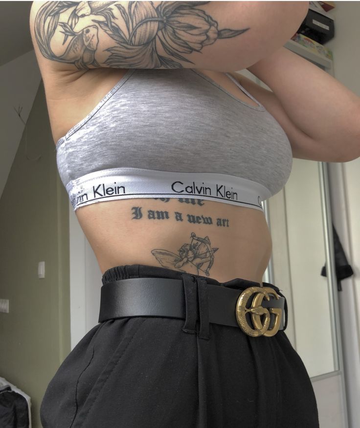 a woman with tattoos on her stomach wearing black pants and a silver top is standing in front of a mirror