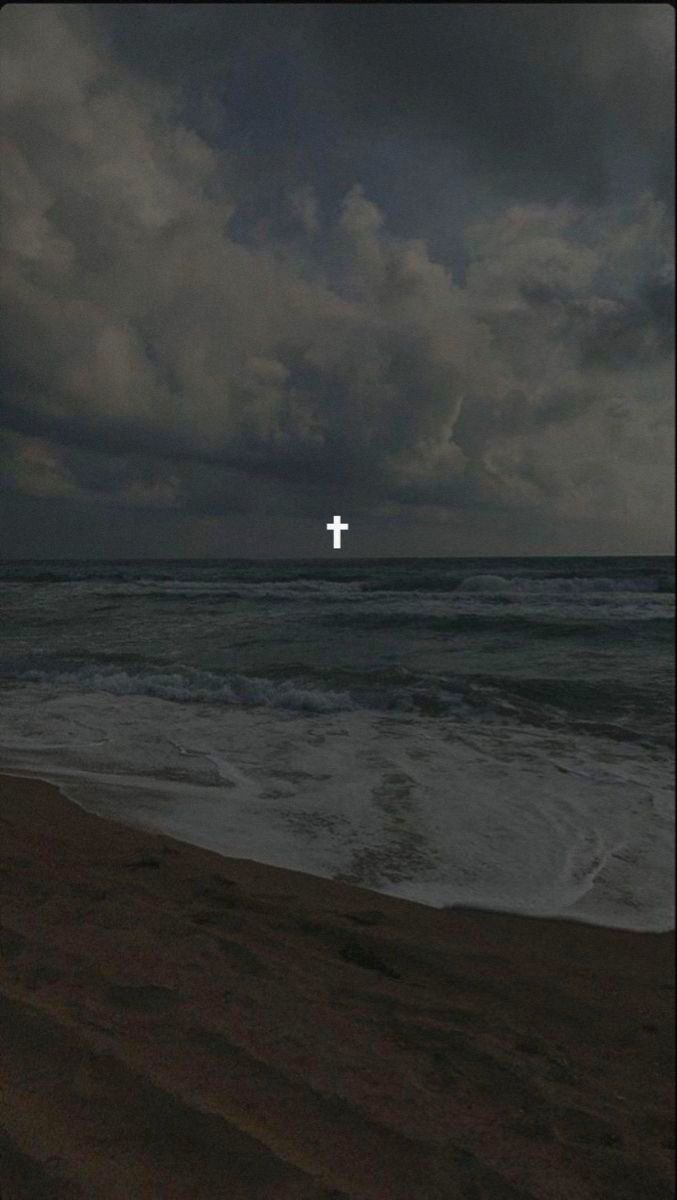 a cross is seen in the distance on an overcast day at the ocean's edge