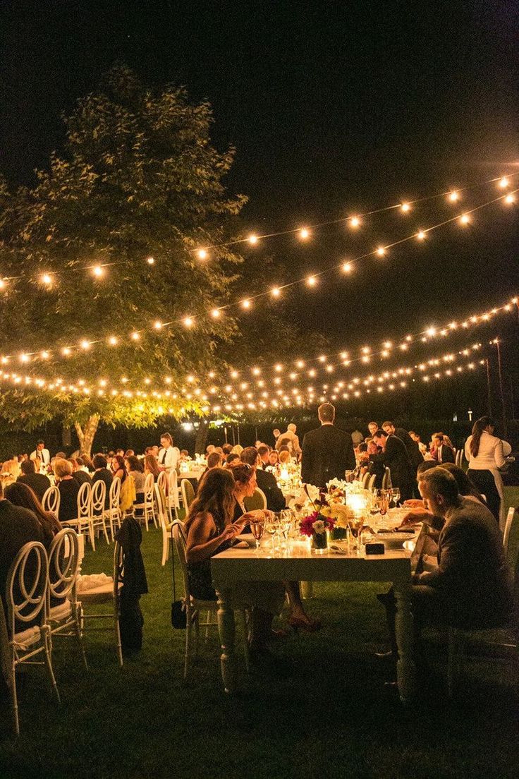 a group of people sitting at tables under string lights in the dark with white chairs