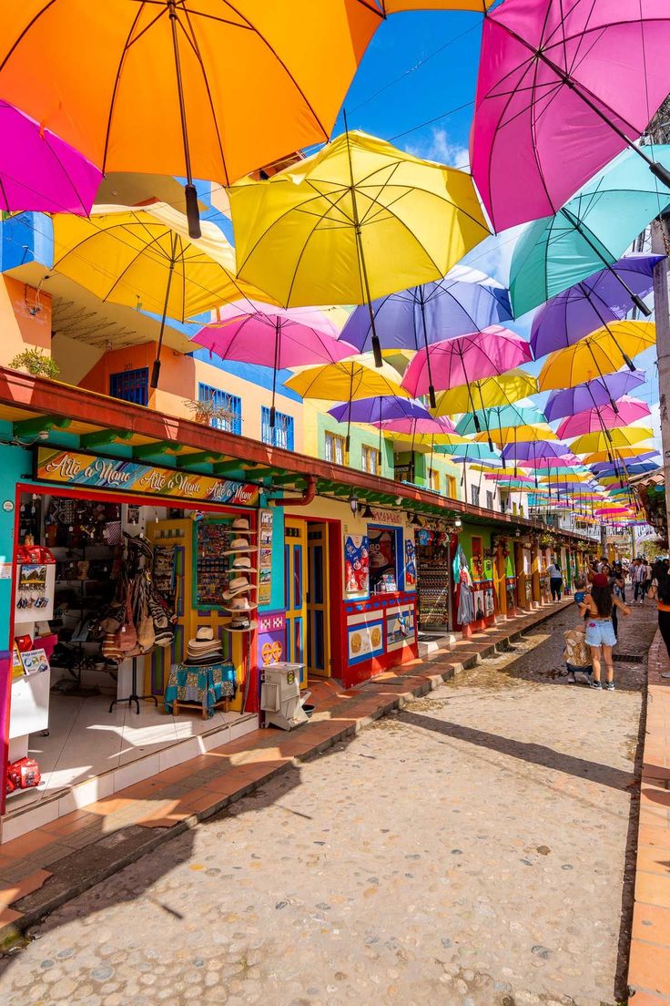 many colorful umbrellas are hanging in the air above a street side market area with people walking by