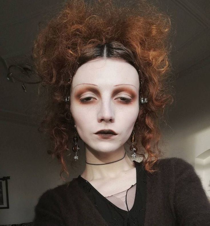 a woman with red hair and white make - up looks at the camera while wearing earrings