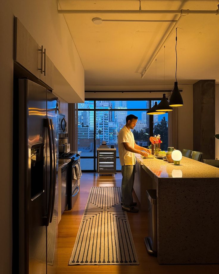 a man preparing food in a kitchen next to a refrigerator and window with city view