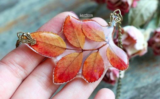 a hand holding a leaf shaped broochle in it's left hand, with pink and red flowers on the other side