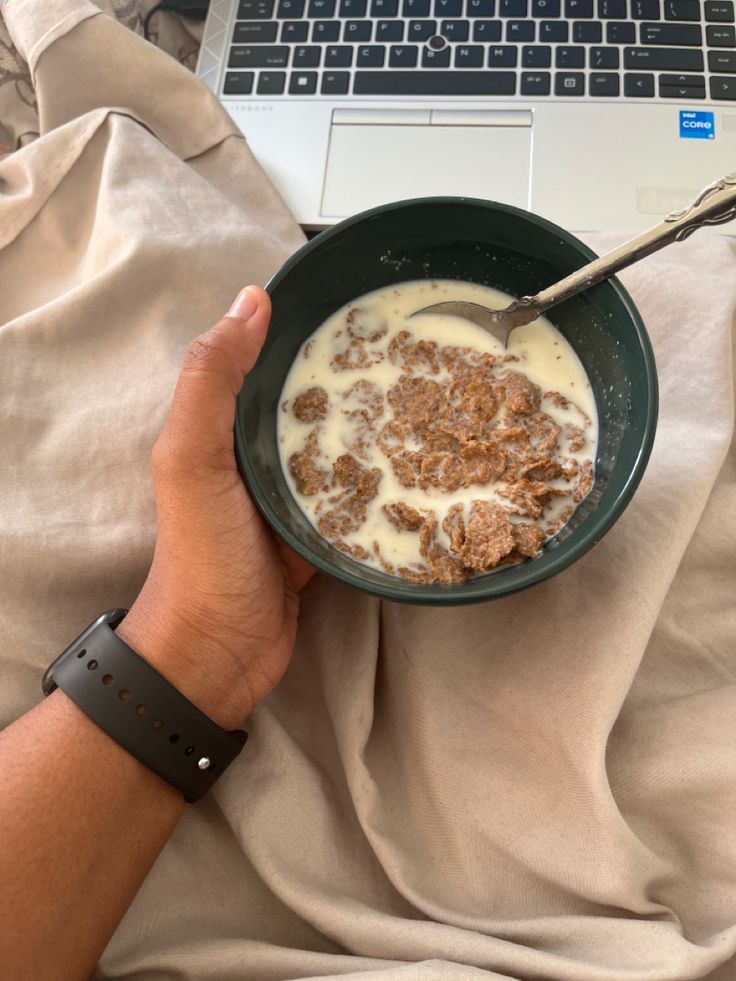 a person is holding a bowl of cereal in front of a laptop on a bed